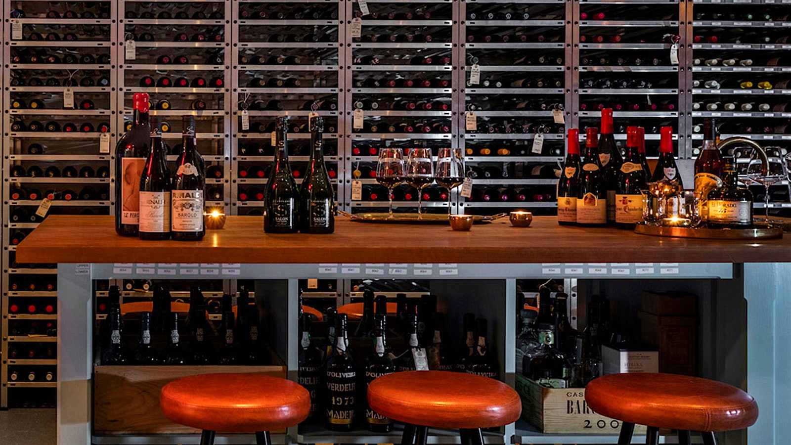  The sleek, modern-design cellar at March, with metal bottle shelves, a table lined with wine bottles and wineglasses and three red, leather stools